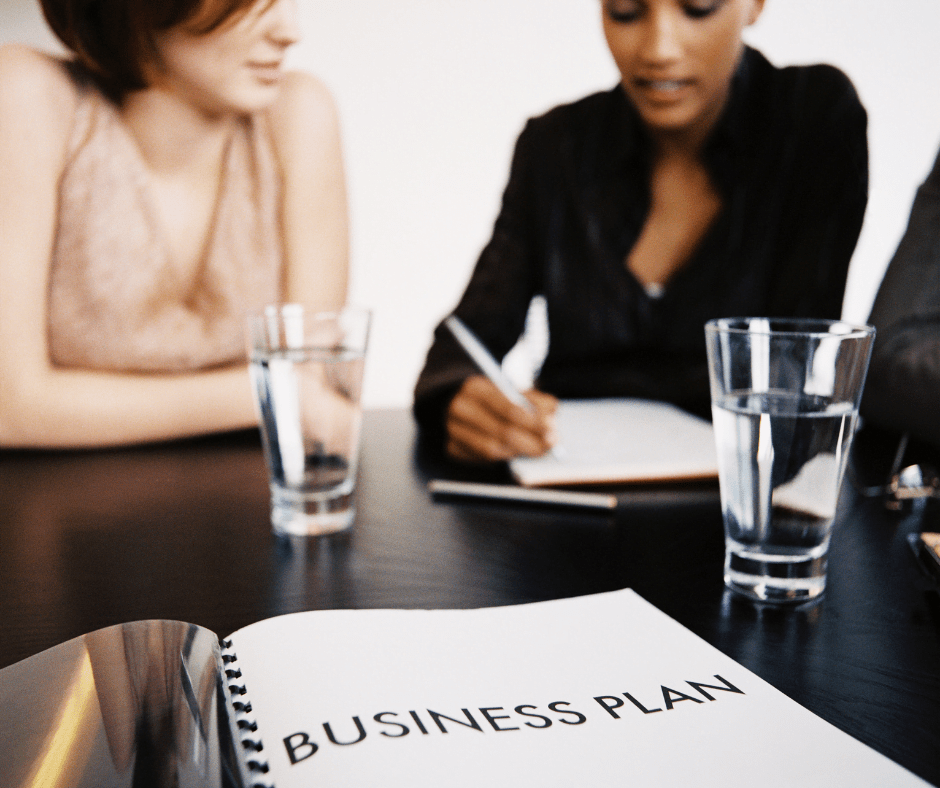 Two women sitting at a desk. One is writing in a note book. There are 2 glasses of water and an open folder with the words BUSINESS PLAN in the foreground of the shot.