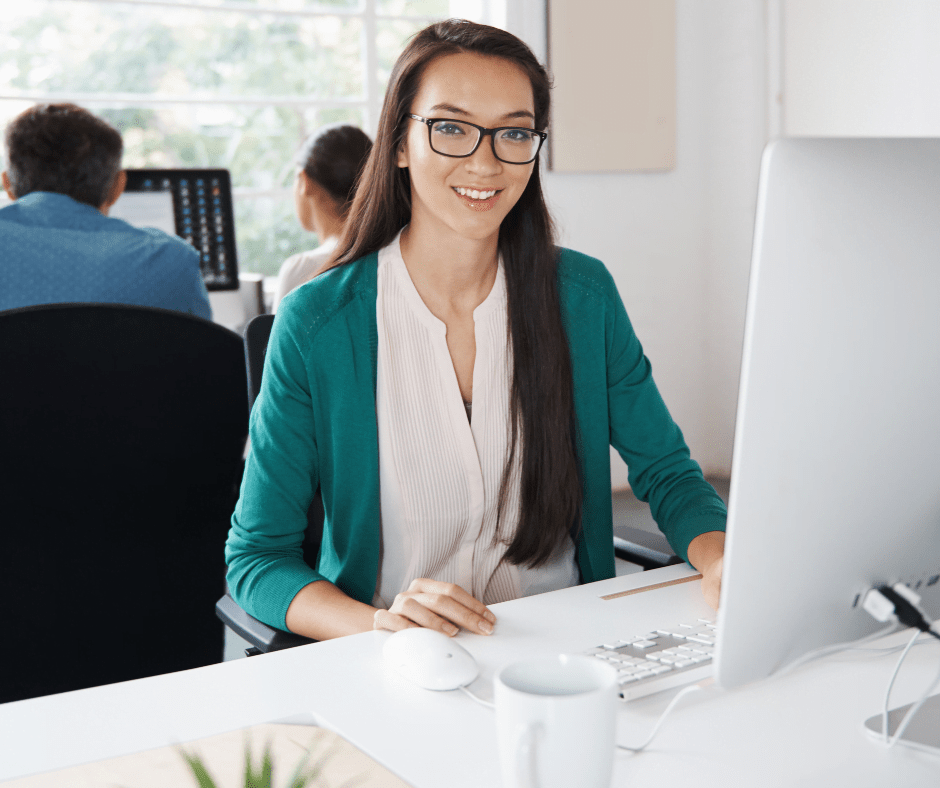 Young woman with long dark hair and glasses, in a green cardigan, sitting at a white desk with a large computer screen. Two colleagues in the background.