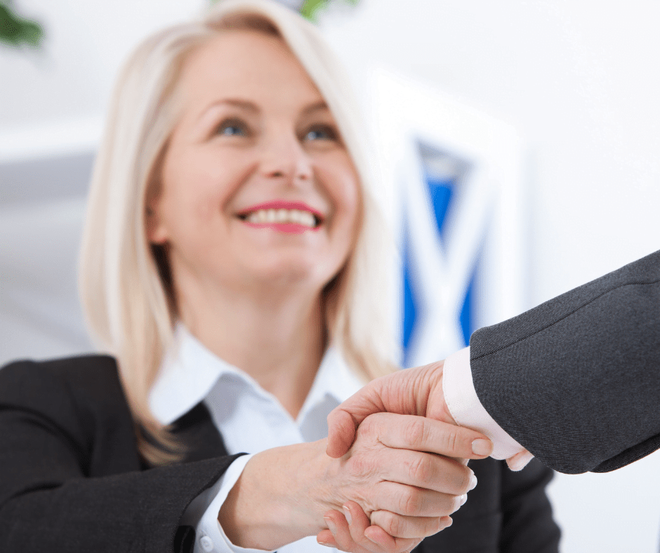 Blonde woman in a black suit, smiling and shaking hands with someone out of shot.