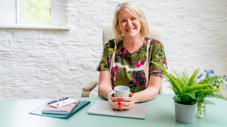 Louise Jenner is sitting at a desk waiting for a career coaching client to arrive. There is a laptop, note books and a plant on the desk. She is holding a mug with both hands.