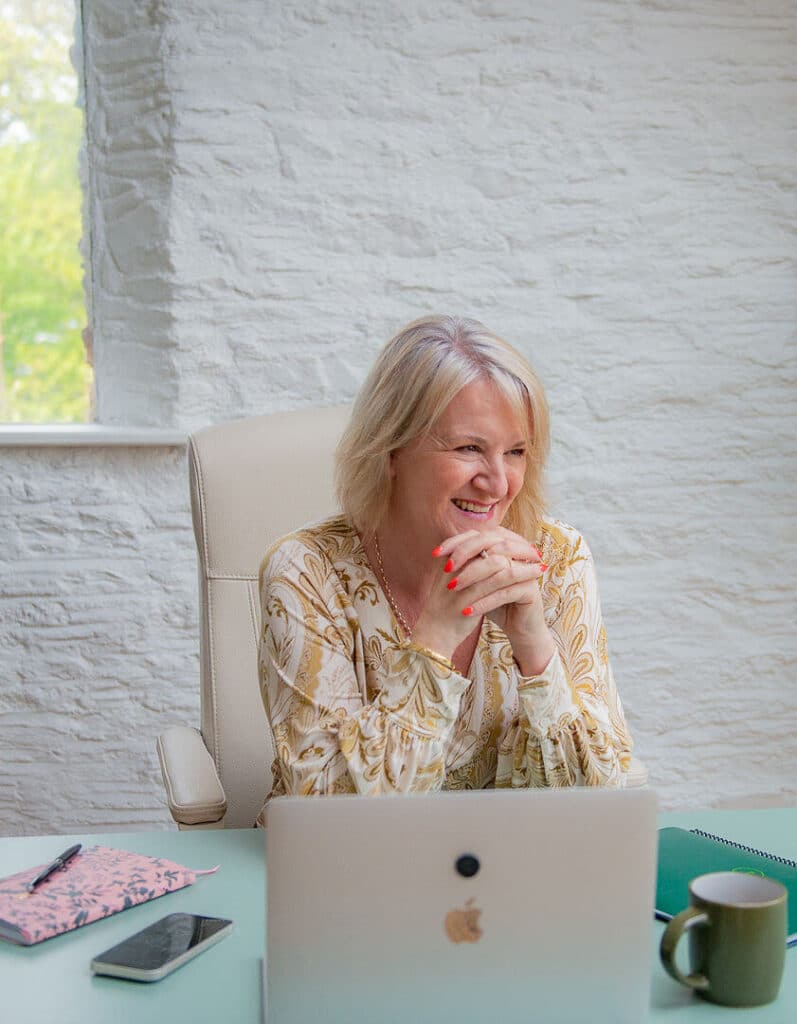 Louise Jenner, The Dream Job Coach is sitting at a desk with her laptop open. She has her head on her clasped hands and is smiling to the right, maybe watching the dream job mindset shift of one of her career coaching clients.