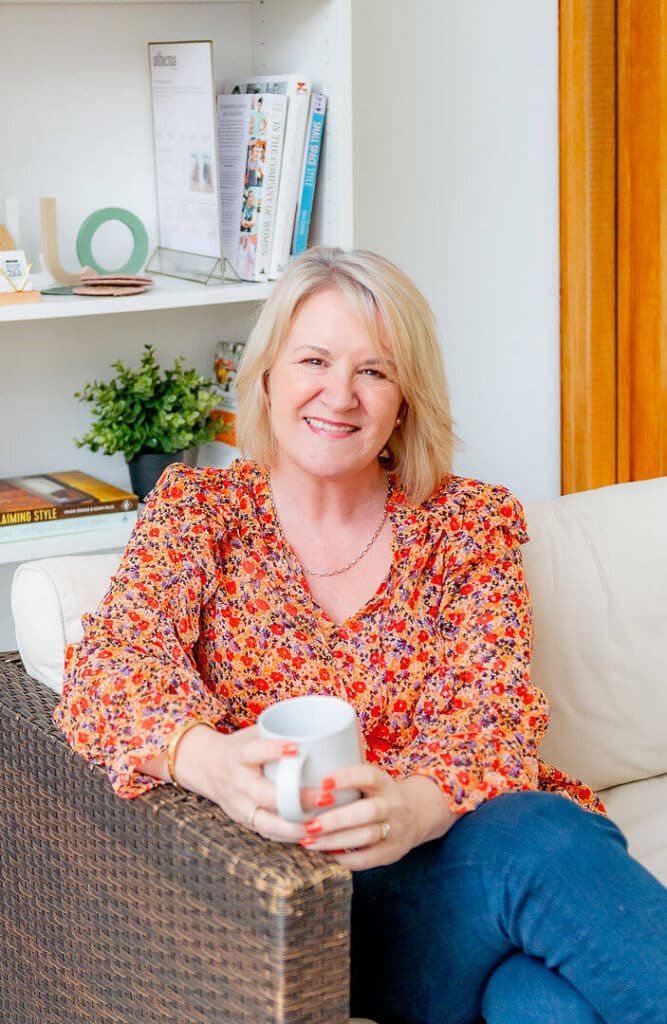 Louise Jenner is sitting on a sofa, wearing an orange floral blouse. She is leaning on the armrest, holding a mug of coffee.