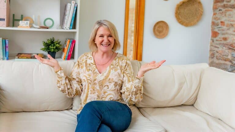 Louise Jenner is sitting on a cream sofa, smiling with her hands up.
